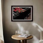 Framed artwork of a car with flowers on a wall above a wooden table with a glass and magazine.