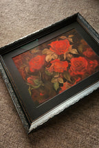 A framed floral art print featuring red roses with green leaves, set in a black frame with a decorative silver inner trim, resting on a textured brown fabric surface.