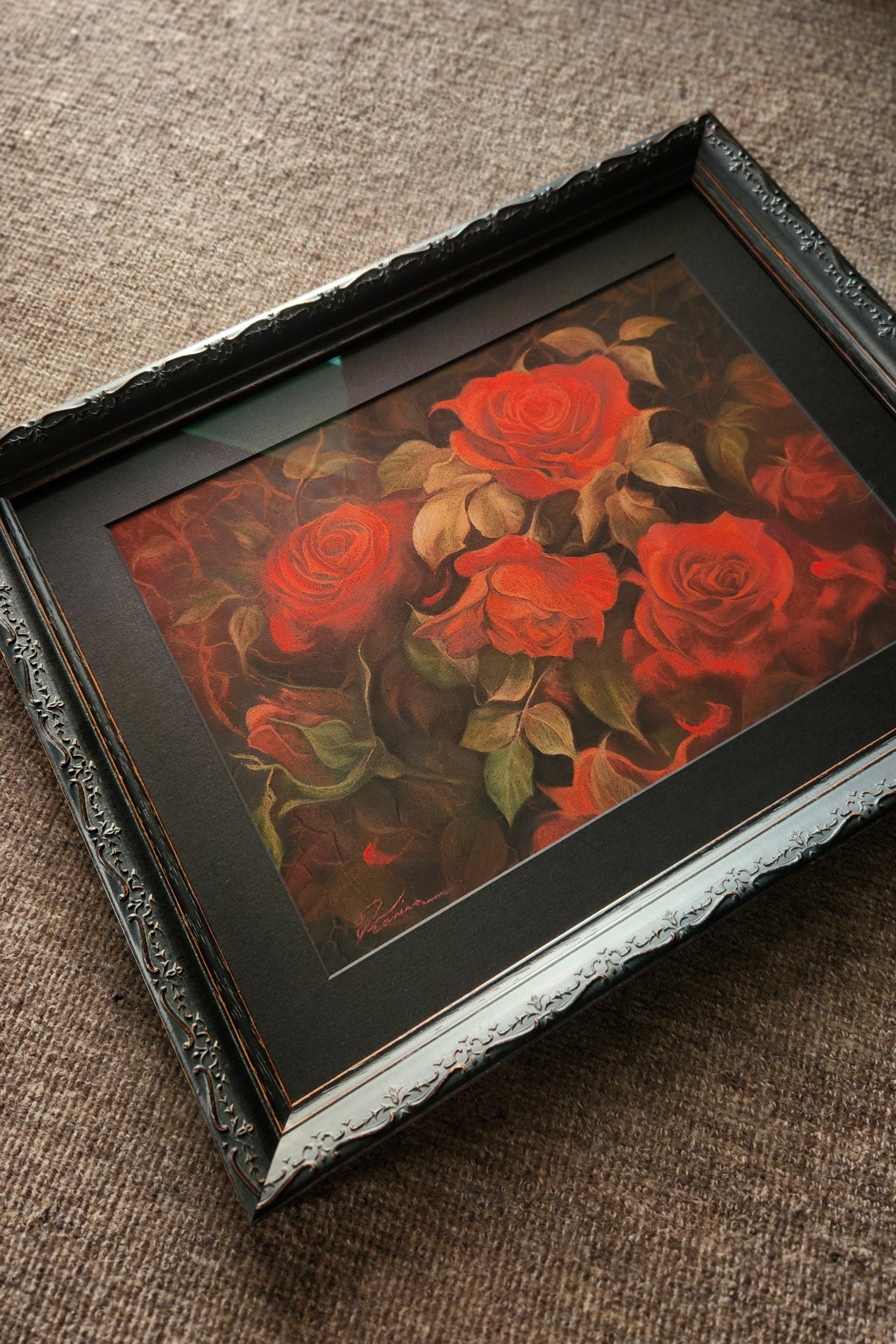A framed floral art print featuring red roses with green leaves, set in a black frame with a decorative silver inner trim, resting on a textured brown fabric surface.