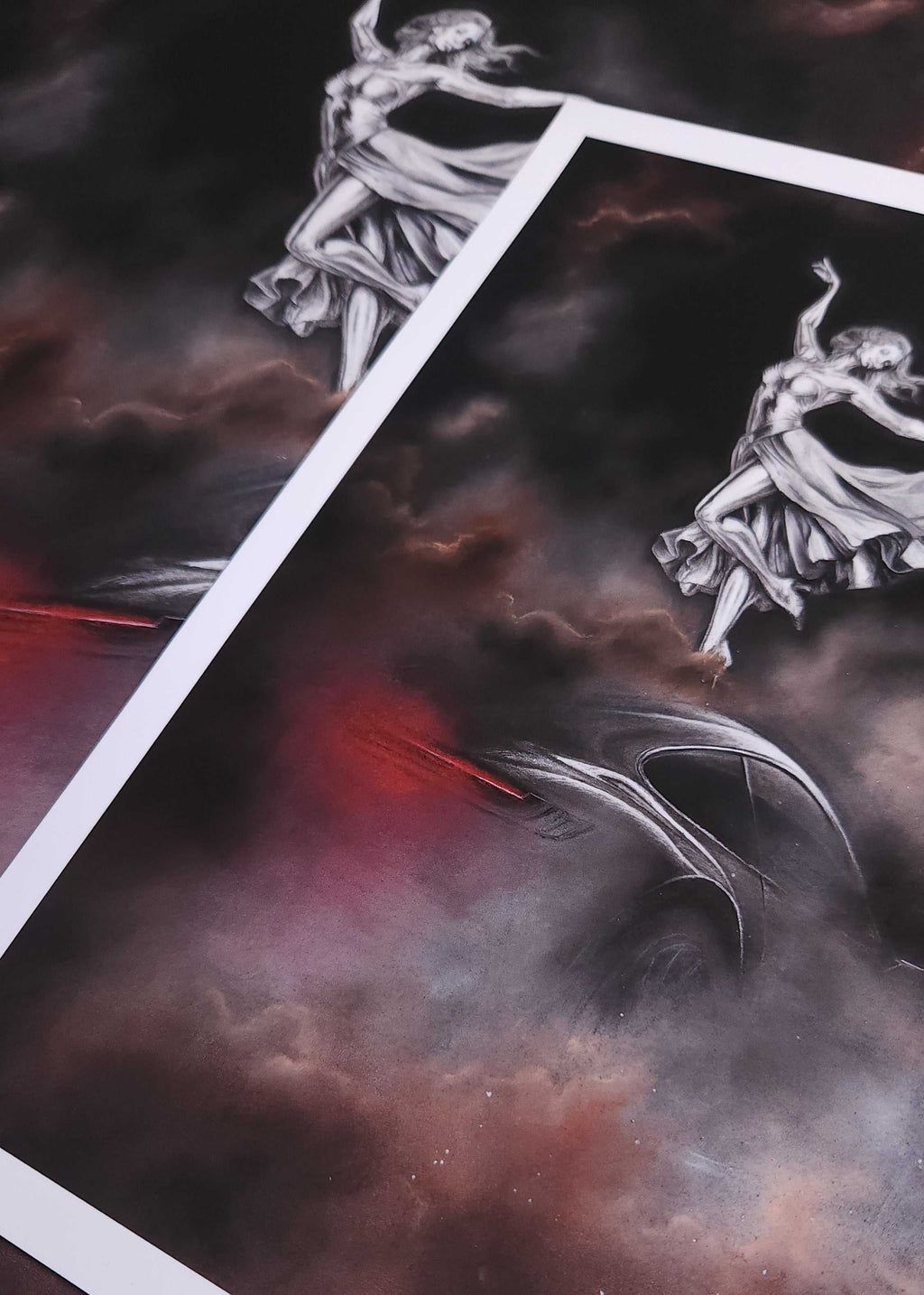 A close-up of a printed artwork or poster showing a dramatic dark scene with a white-bordered sheet laid over another matching print. The artwork features a grayscale dancing female figure against smoky clouds with red and brown undertones, plus a dark curved object in the lower area.