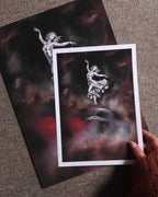 A hand holds a white-bordered art print showing a classical statue-like figure floating in a dark smoky scene, with a larger matching print laid behind it on a carpeted floor.
