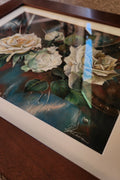 A framed floral art print featuring cream and white roses with green leaves on a dark blue and brown background, shown resting on a wooden surface.