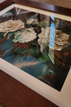 A framed floral art print featuring cream and white roses with green leaves on a dark blue and brown background, shown resting on a wooden surface.