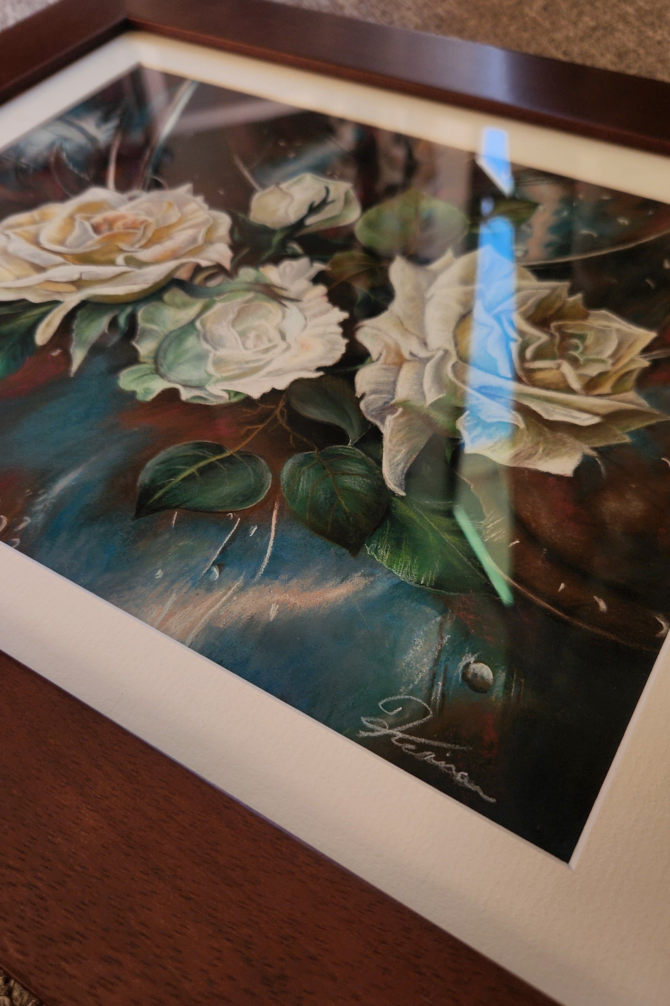 A framed floral art print featuring cream and white roses with green leaves on a dark blue and brown background, shown resting on a wooden surface.