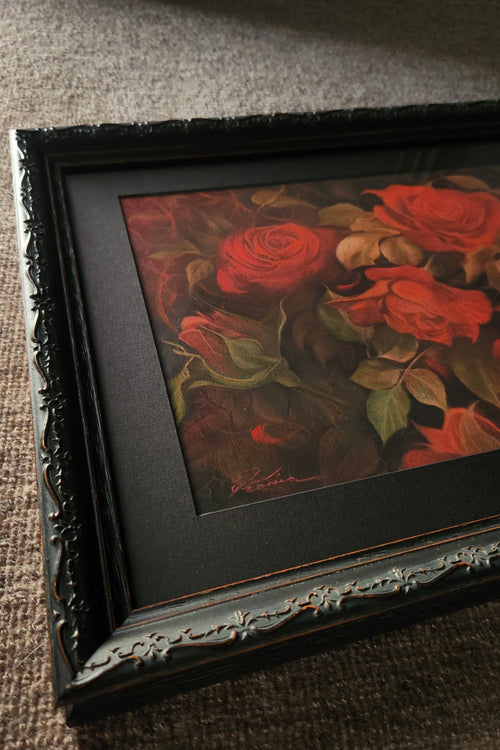 A framed floral art print featuring red roses with green leaves, set in a black ornate frame and resting on a textured gray carpet.