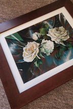 Framed floral artwork showing three cream and pale green roses with green leaves on a dark blue and brown abstract background, set in a white mat and dark brown wooden frame on a textured beige surface.